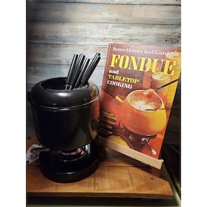 May include: A black fondue pot with fondue forks and a cookbook titled "Fondue and Tabletop Cooking" by Better Homes and Gardens. The cookbook is propped up on a wooden stand. The image is set against a wood-paneled background.