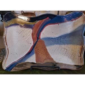 May include: Rectangular ceramic serving platter with a wavy edge. The platter features a white base with blue, red, and brown abstract designs. The design includes embossed leaf patterns and is displayed on a black metal stand.
