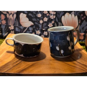 May include: Two ceramic mugs on a wooden tray. One mug is a dark blue with white floral accents, and the other is white with black abstract shapes. Both have handles and are suitable for hot beverages.