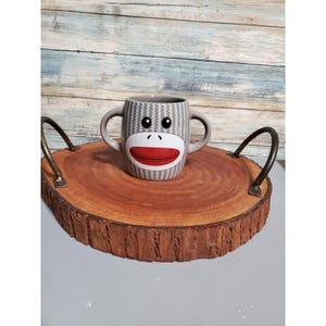 May include: A gray and white patterned mug with a monkey face design, featuring a red mouth and black eyes, sits on a round wooden serving tray with metal handles. The tray has a natural bark edge and a light brown surface.