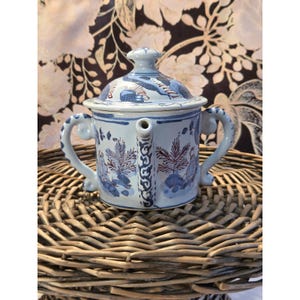 May include: A blue and white ceramic teapot with a lid, spout, and two handles. The teapot features a floral and foliate design with red and blue accents. It is sitting on a woven wicker surface.