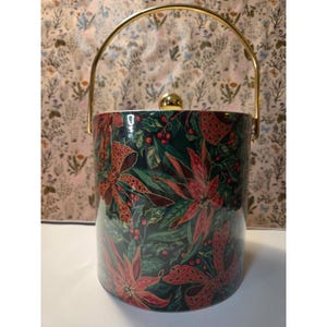 May include: A ceramic ice bucket with a gold handle and lid. The bucket is cylindrical and decorated with a festive pattern of red poinsettias, green leaves, and red berries on a dark green background. The handle is arched.