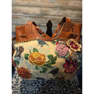 May include: A large tote bag with a floral and bird pattern. The bag features brown leather handles and trim, with crocheted flower embellishments. The main fabric is a cream color with colorful floral and bird designs.