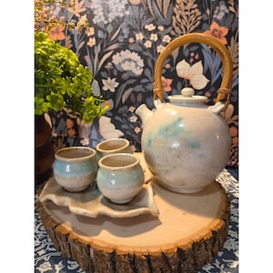 May include: A ceramic tea set with a teapot and three small cups, all in a cream color with blue and green accents. The teapot has a bamboo handle. The cups sit on a leaf-shaped tray, all displayed on a wooden slab.