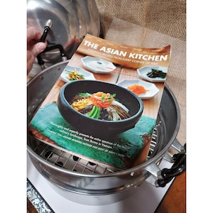 May include: A stainless steel steamer with a cookbook titled "The Asian Kitchen" open inside. The cookbook features a photo of a black bowl filled with colorful Asian cuisine. The steamer has a black handle and a lid.
