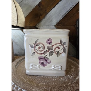 May include: A square, cream-colored ceramic planter with a scalloped top edge and a raised decorative base. The front features a painted design of two purple roses with green leaves and brown swirling vine accents on a smooth, glossy surface.