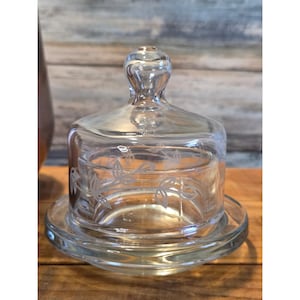 May include: A clear glass butter dish with a domed lid and etched floral design. The dish sits on a round base and has a small handle on top of the lid. The glass is transparent, allowing the contents to be visible.