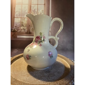 May include: Cream-colored ceramic pitcher with a bulbous base and a slender neck. The pitcher features a curved handle and is adorned with pink and red floral patterns. A small lid sits atop the pitcher. The pitcher is displayed on a wooden tray.