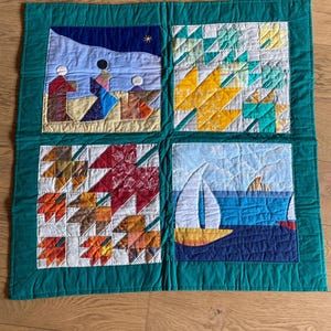 Peut inclure: Une courtepointe faite à la main avec quatre panneaux carrés. Chaque panneau présente un motif différent : une scène de nativité, des motifs géométriques jaunes et verts, des feuilles d'automne et un voilier sur la mer. La courtepointe est bordée d'un tissu vert.