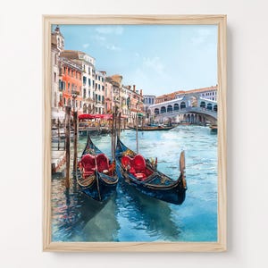 May include: Watercolor painting of Venice, Italy, featuring two gondolas with red seating, floating on blue water. The artwork showcases colorful buildings, a bridge, and a clear blue sky, all framed in a light wood frame.