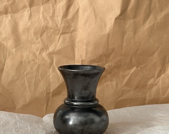 Può includere: Un piccolo vaso grigio scuro con una base bulbosa e una parte superiore svasata. Il vaso ha una finitura liscia e metallica e una forma classica. Si trova su una superficie bianca contro uno sfondo di carta marrone. Il vaso è probabilmente in metallo.