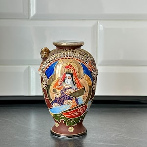 Könnte beinhalten: Kleine, verzierte Vase mit braunem Sockel und goldenem Rand. Die Vase zeigt eine detaillierte Illustration einer Figur mit Heiligenschein, umgeben von goldenen, blauen und roten Akzenten. An der Seite befindet sich ein kleiner Griff.
