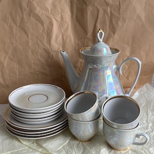 Puede incluir: Un juego de té nacarado con ribete dorado. El juego incluye una tetera, platillos y tazas de té. Los platillos y las tazas de té están apilados. La tetera tiene un asa dorada y una tapa con un adorno dorado. El juego de té está sobre una superficie blanca.
