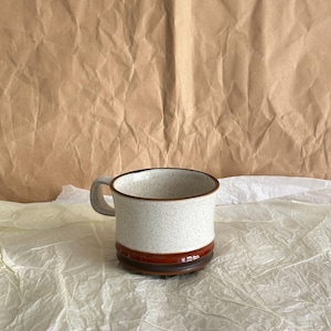 Puede incluir: Una taza de cerámica con cuerpo blanco moteado, borde marrón y una banda marrón y roja cerca de la base. La taza tiene un asa gris y está sobre un fondo neutro.