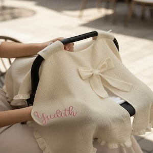 Könnte beinhalten: Cremefarbene Baby-Autositzabdeckung mit Schleife und Rüschenkanten. Der Bezug hat den Namen "Judith" in rosa Schrift gestickt. Der Stoff scheint ein weiches, strukturiertes Material zu sein.