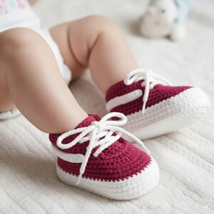 May include: A pair of handmade baby sneakers in a deep red color with white accents. The crochet shoes have white laces and a white sole. The sneakers are designed to look like miniature versions of adult sneakers.