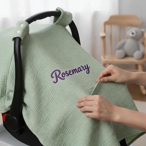 Könnte beinhalten: Eine hellgrüne Baby-Autositzabdeckung mit dem Namen "Rosemary" in lila Schrift gestickt. Die Abdeckung besteht aus einem strukturierten Stoff und ist über einem schwarzen Autositzgriff drapiert. Hände passen die Abdeckung an.