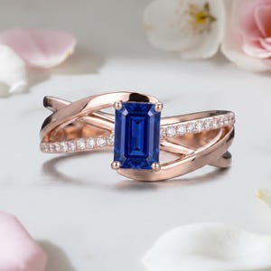 May include: A rose gold ring featuring a central emerald-cut blue sapphire held by four prongs. The band consists of two intertwining, polished rose gold strands, with one strand accented by a row of small, clear, round-cut pavé-set white stones.