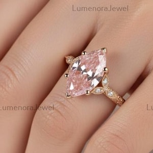Könnte beinhalten: Ein roségoldener Ring mit einem großen, marquiseförmigen rosa Edelstein. Das Band ist mit kleinen, filigranen Verzierungen versehen. Der Ring wird an einer Hand gezeigt, um das Design und die Farbe des Schmucks hervorzuheben.