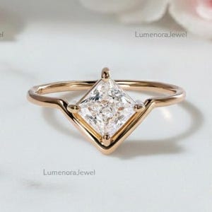 May include: A gold ring with a princess-cut diamond center stone. The diamond is set in a V-shaped setting. The ring is on a white surface with flowers in the background. The text "LumenoraJewel" is visible.