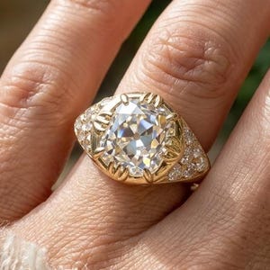 Op de afbeelding: Een gouden ring met een grote, kussengeslepen middensteen vastgehouden door klauwen, geflankeerd door kleinere ronde accentstenen in de band. De metalen band heeft een gepolijste afwerking en een breed, taps toelopend ontwerp.