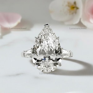 Puede incluir: Un anillo de compromiso de diamantes con una gran piedra central en forma de pera y dos diamantes baguette más pequeños a cada lado. El anillo está engastado en una banda plateada. El fondo incluye pétalos de flores blancos y rosas.