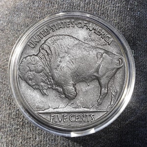 May include: A silver-colored coin featuring a detailed depiction of a bison. The coin is encased in a clear protective holder. The text "UNITED STATES OF AMERICA" and "FIVE CENTS" are visible.