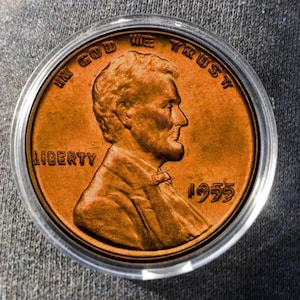 May include: A copper-coloured 1955 Lincoln penny in a clear plastic case. The coin shows a profile of Abraham Lincoln, with the inscriptions "IN GOD WE TRUST" and "LIBERTY". The year "1955" is also displayed on the coin.