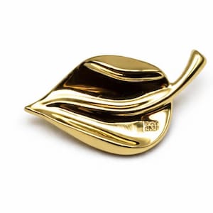 May include: A gold-colored leaf-shaped brooch. The brooch has a smooth, reflective surface and a stem detail. The leaf is slightly curved, with visible veins. The brooch is set against a white background.