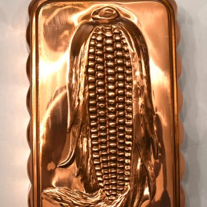 May include: A copper-colored, rectangular mold with a detailed embossed design of an ear of corn. The mold has a scalloped edge and a small brass ring for hanging. The corn design is highly detailed, with individual kernels and husks.