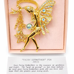 Könnte beinhalten: Ein goldfarbener Feen-Anstecknadel mit schillernden Kristallakzenten. Die Fee hält eine Kristallkugel und einen Sternanhänger. Die Brosche wird in einer rosa Schachtel mit einer bedruckten Karte mit der Aufschrift "FAIRY GODMOTHER" PIN präsentiert.