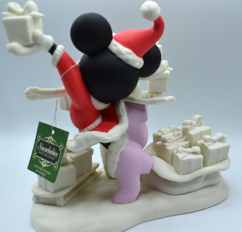 Op de afbeelding: Een Snowbabies-beeldje van Mickey Mouse in een kerstmanpak, met cadeaus. Het beeldje is voornamelijk wit, met rode en zwarte accenten. Het label luidt "Snowbabies". Het beeldje staat op een witte basis met andere cadeaus.