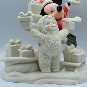 Op de afbeelding: Een feestelijk beeldje met een sneeuwpop in een hooded jumpsuit die twee cadeau dozen vasthoudt. Mickey Mouse met een kerstmuts staat op de achtergrond. Verschillende andere cadeau dozen staan op een met sneeuw bedekte basis. Het beeldje is voornamelijk gebroken wit.