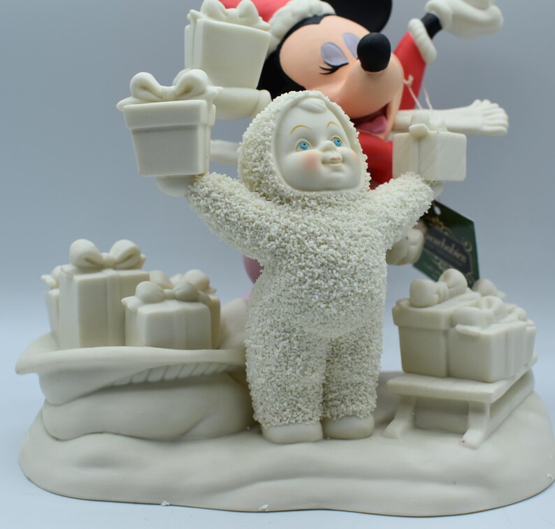 Op de afbeelding: Een feestelijk beeldje met een sneeuwpop in een hooded jumpsuit die twee cadeau dozen vasthoudt. Mickey Mouse met een kerstmuts staat op de achtergrond. Verschillende andere cadeau dozen staan op een met sneeuw bedekte basis. Het beeldje is voornamelijk gebroken wit.