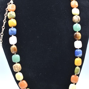 May include: A multi-coloured gemstone necklace with square beads. The necklace is made of various stones, including tiger's eye, lapis lazuli, and aventurine. The necklace is displayed on a black velvet mannequin.