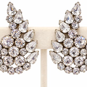 May include: A pair of statement earrings featuring a cluster of clear, faceted, round and pear-shaped crystals set in a silver-toned metal frame. The design is asymmetrical, with a curved, wing-like silhouette that tapers toward the top.