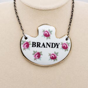 May include: A white ceramic pendant necklace featuring a gold-toned rim and a floral pattern of small pink roses. The word BRANDY is printed in black capital letters across the center of the pendant, which hangs from a dark metal chain.