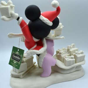 Op de afbeelding: Een Snowbabies-beeldje van Mickey Mouse in een kerstmanpak, met cadeaus. Het beeldje is voornamelijk wit, met rode en zwarte accenten. Het label luidt "Snowbabies". Het beeldje staat op een witte basis met andere cadeaus.
