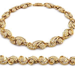May include: A gold-toned necklace and bracelet set. The jewelry features a repeating swirl design, each element encrusted with small, sparkling gemstones. The necklace is clasped, and the bracelet is laid out below it.