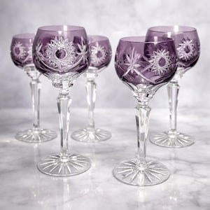 May include: Five purple crystal wine glasses with starburst designs. The glasses have a clear stem and base, and the bowl is a deep purple color. The starburst design is etched into the bowl and base of the glasses. The glasses are arranged on a marble surface.
