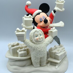 Op de afbeelding: Een kerstbeeldje met Mickey Mouse in een kerstmuts en rode jas, met cadeaus. Een sneeuwman-achtig figuur met een capuchonjas houdt ook cadeaus vast. De basis omvat een slee en een tas vol cadeaus, allemaal in een cr&egrave;mekleur.