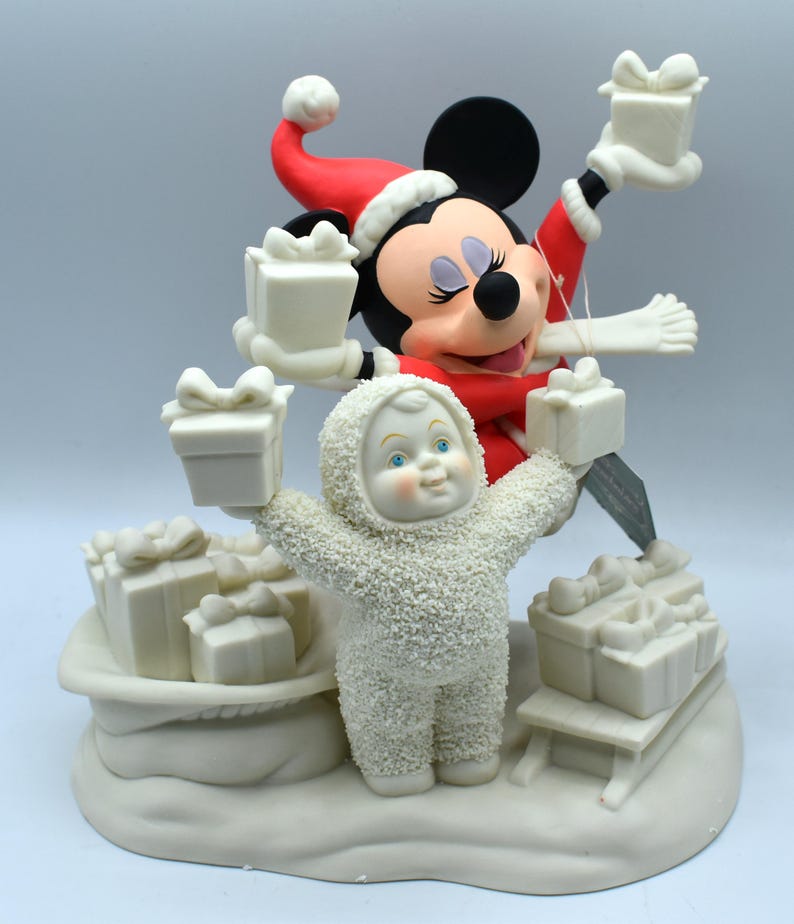 Op de afbeelding: Een kerstbeeldje met Mickey Mouse in een kerstmuts en rode jas, met cadeaus. Een sneeuwman-achtig figuur met een capuchonjas houdt ook cadeaus vast. De basis omvat een slee en een tas vol cadeaus, allemaal in een cr&egrave;mekleur.