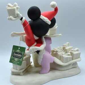 Op de afbeelding: Een Snowbabies Disney Collection beeldje met Mickey Mouse in een kerstmanpak, met een cadeau. Het beeldje is wit met rode en zwarte accenten, en bevat een label met "Look What We Have For Mickey!"
