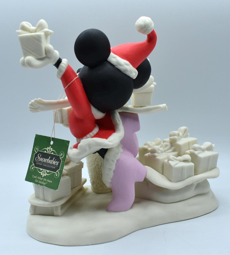 Op de afbeelding: Een Snowbabies Disney Collection beeldje met Mickey Mouse in een kerstmanpak, met een cadeau. Het beeldje is wit met rode en zwarte accenten, en bevat een label met "Look What We Have For Mickey!"