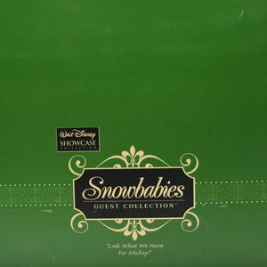 Op de afbeelding: Een groene doos met het logo "Walt Disney Showcase Collection". De doos heeft het logo "Snowbabies Guest Collection" in een decoratief kader, met de tekst "Look What We Have For Mickey!"