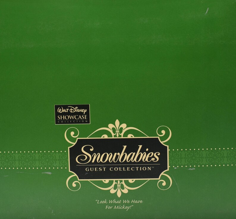 Op de afbeelding: Een groene doos met het logo "Walt Disney Showcase Collection". De doos heeft het logo "Snowbabies Guest Collection" in een decoratief kader, met de tekst "Look What We Have For Mickey!"