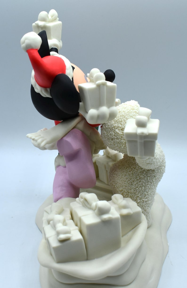 Op de afbeelding: Een beeldje met een stripfiguur met een kerstmuts, zwarte oren en roze kleding, met witte geschenkdozen. Er is ook een sneeuwpopfiguur aanwezig, met meer geschenkdozen op een besneeuwde basis. Een feestelijke kerstversiering.