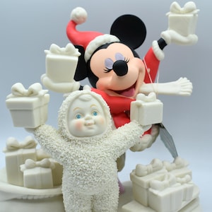 Op de afbeelding: Een feestelijk beeldje met Mickey Mouse in een kerstmuts en rode jas, met een cadeau. Een sneeuwpopfiguur in een wit getextureerd pak houdt ook een cadeau vast. Verschillende witte geschenkdozen.