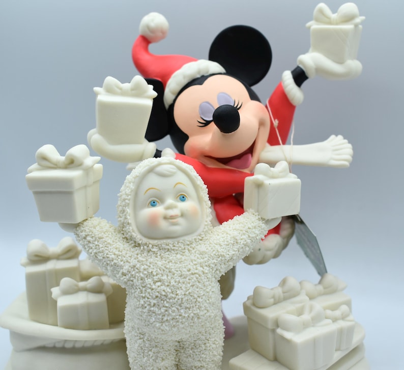 Op de afbeelding: Een feestelijk beeldje met Mickey Mouse in een kerstmuts en rode jas, met een cadeau. Een sneeuwpopfiguur in een wit getextureerd pak houdt ook een cadeau vast. Verschillende witte geschenkdozen.