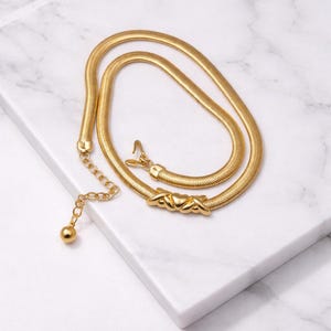 May include: A gold-colored belt with a snake chain design. The belt features a hook closure and a decorative section with intertwined shapes. A small chain with a ball charm hangs from the belt.
